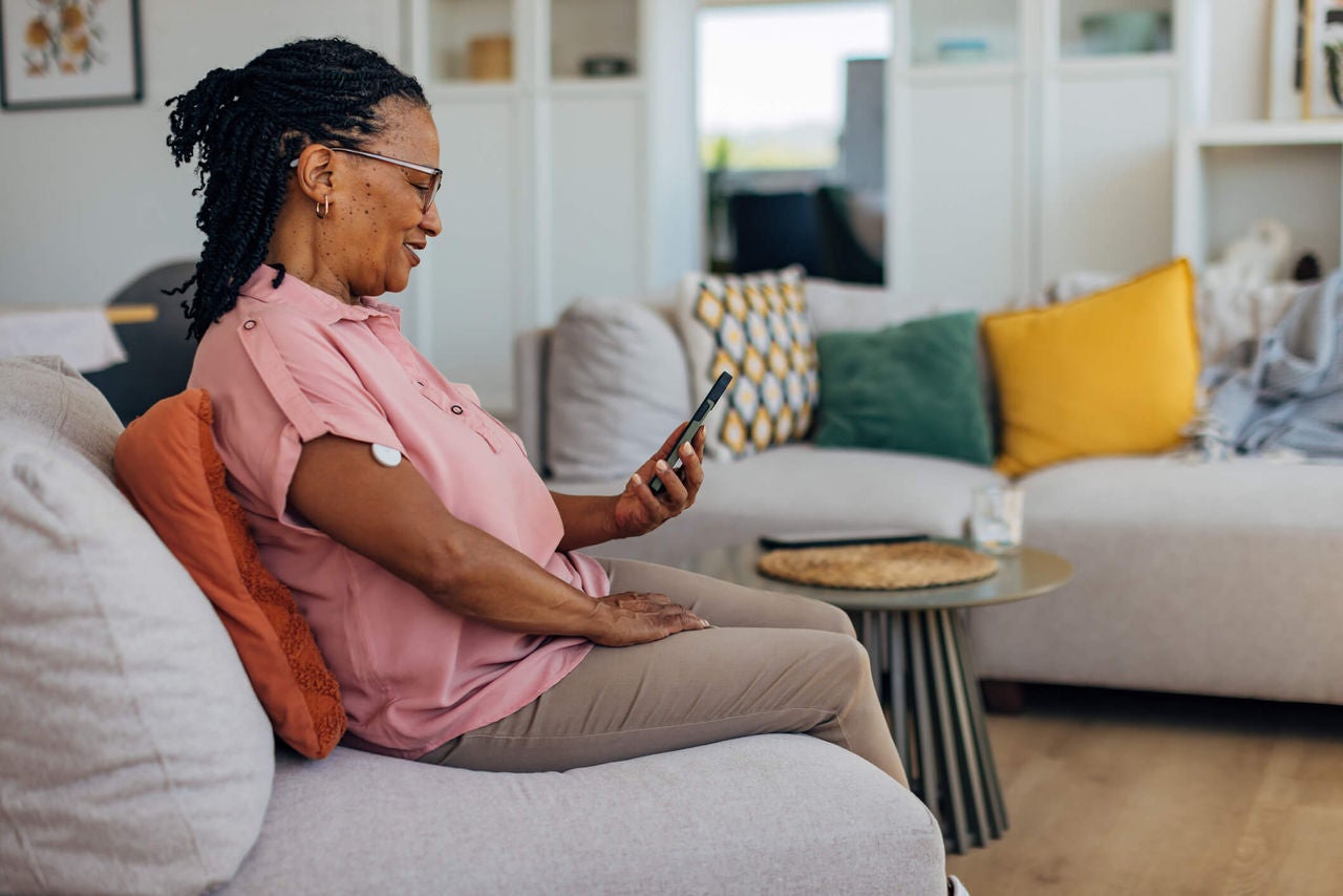 Woman wearing a continuous glucose monitor on her arm, sitting on a couch looking at a smart phone while smiling. 