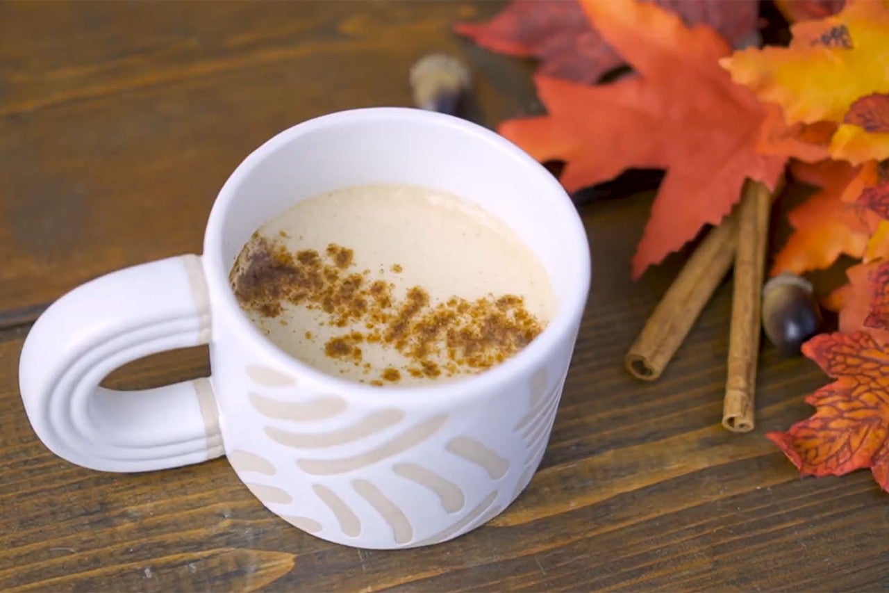 A white mug filled with a creamy beverage, topped with crumbled toppings, surrounded by autumn leaves and cinnamon sticks on a wooden surface.