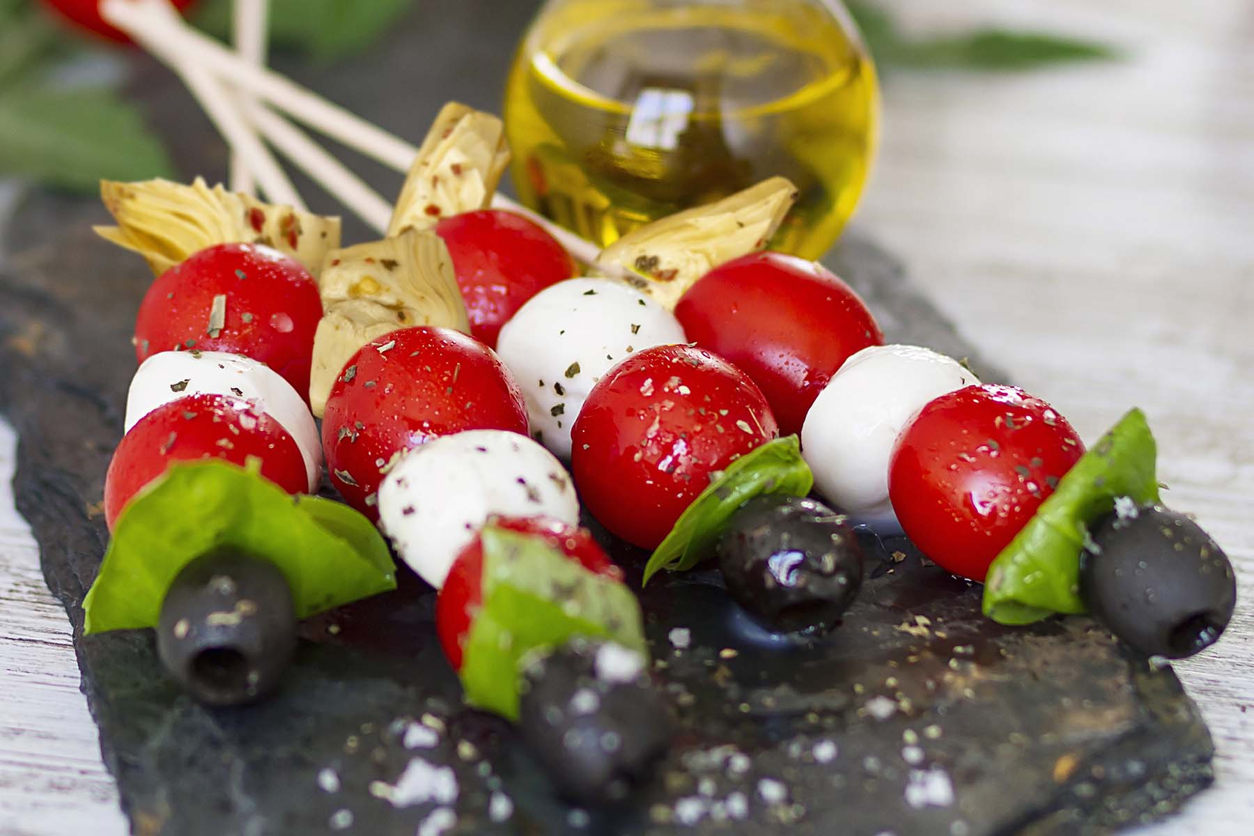 Skewers of cherry tomatoes, mozzarella balls, and fresh basil, interspersed with black olives, arranged on a slate platter, with a small bottle of olive oil in the background.