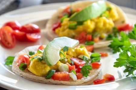 Two breakfast tacos on a white plate. The white corn tortillas are filled with scrambled eggs, tomato, peppers, green onion, cheese, cilantro and avocado. The lighting is very bright, and the colors are vibrant. Focus is on the taco in the foreground and the depth of field is very shallow.