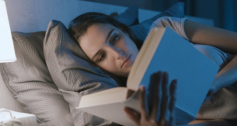 Young woman lying in bed, reading a book under soft bedside lamp light, with a calm and focused expression.