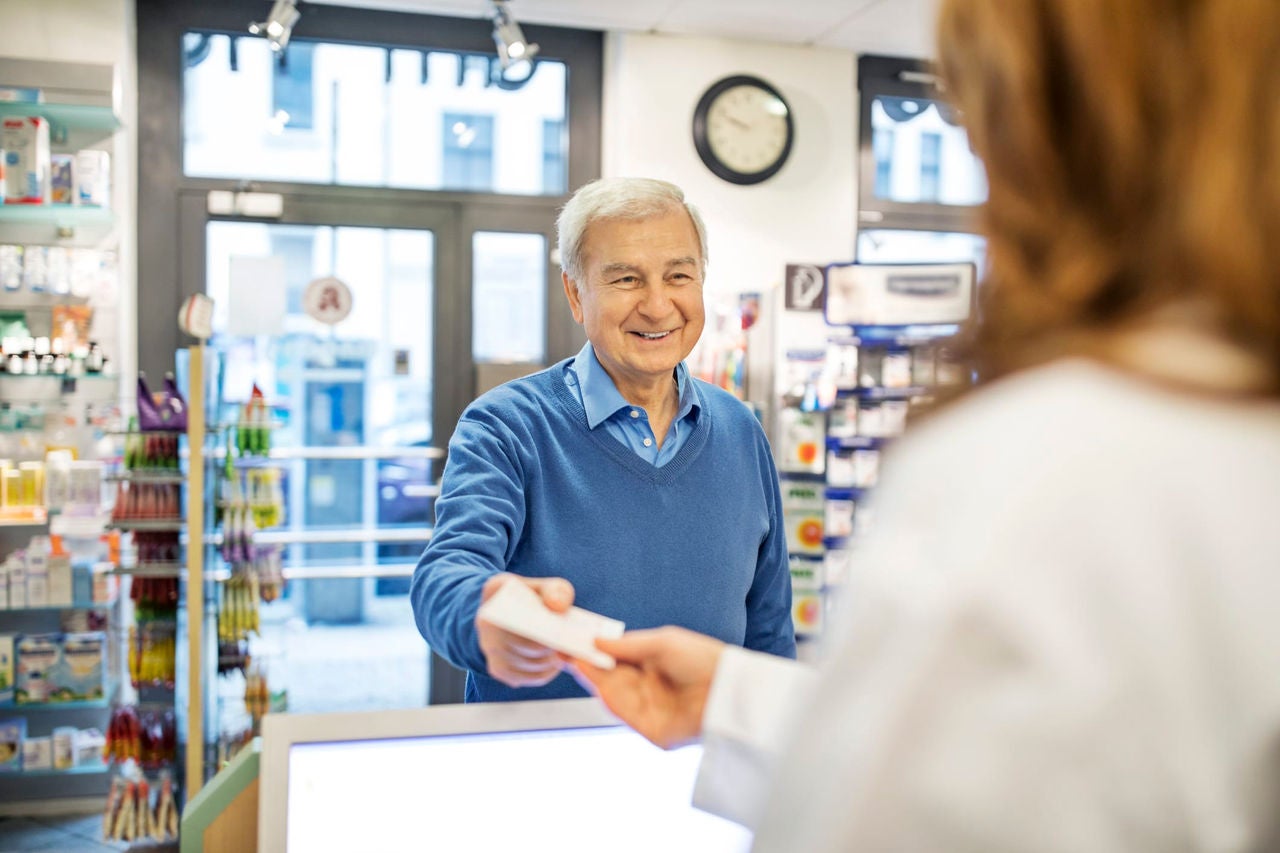 Smiling senior man giving prescription to female pharmacist in store. Chemist is assisting male customer at brightly lit pharmacy. They are standing at checkout counter.
