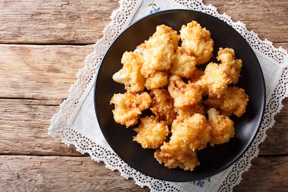 A black plate with fried cauliflower florets, placed on a lace doily atop a rustic wooden table.