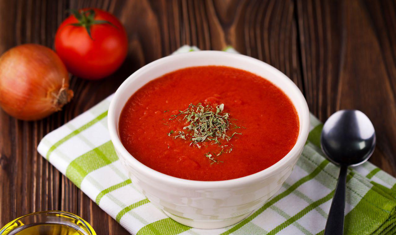 Tomato soup in a white bowl sitting on a green and white towel with a spoon next to it and an onion and tomato in the background