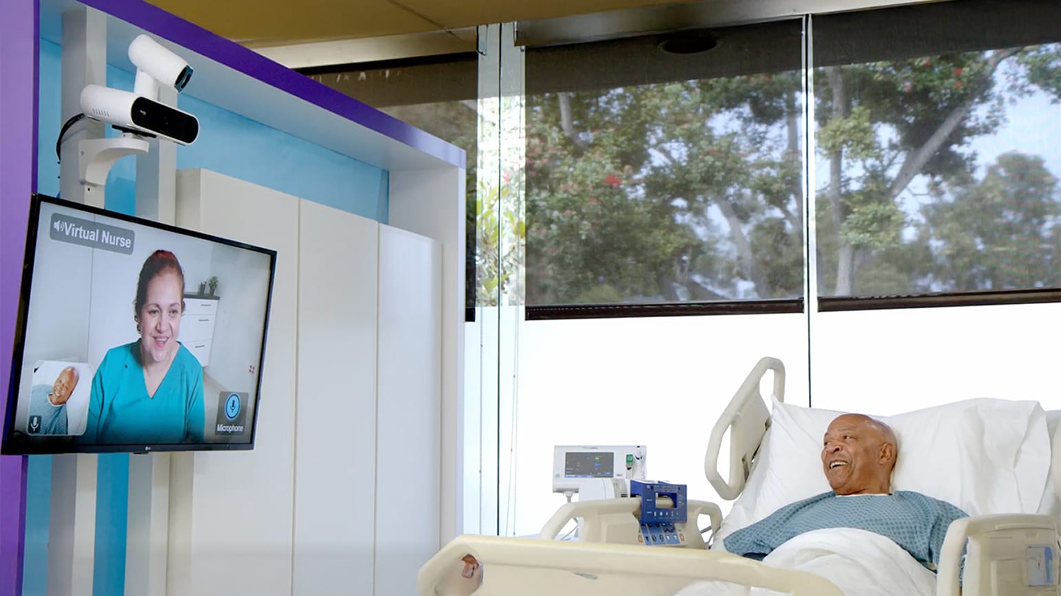 Patient in a hospital bed smiling at a wall-mounted screen showing a virtual nurse on a video call.