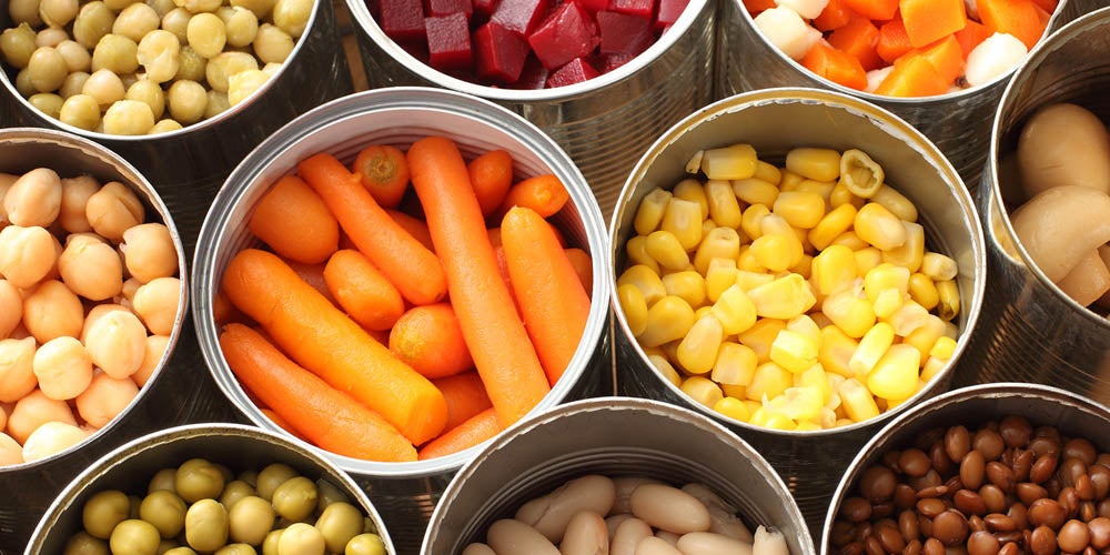 Variedad de vegetales y frutas enlatadas ordenadas en latas individuales, con colores vivos como zanahorias, maíz y porotos mixtos.