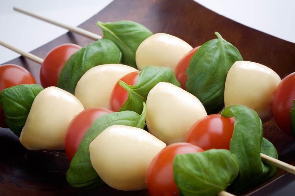 Brochetas de tomates cherry, bolitas de mozzarella y hojas de albahaca fresca dispuestas en un plato oscuro, mostrando un aperitivo colorido y fresco