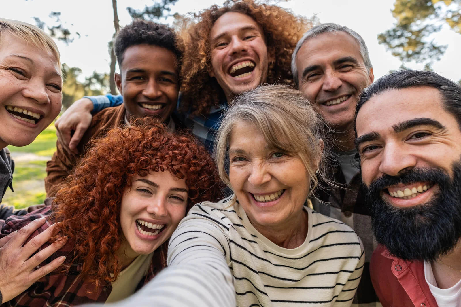 Smiling diverse group of adults taking a close-up selfie outdoors, standing together and looking at the camera.