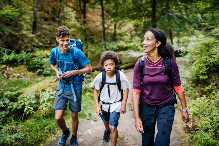 Women with her two children wearing athletic clothing and hiking backpacks smiling while hiking on a woodland trail.