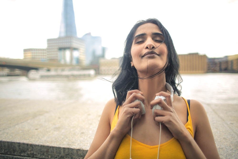 A woman in a yellow top holding the headphones that are around her neck with buildings in the background. Her eyes are closed in a peaceful expression.