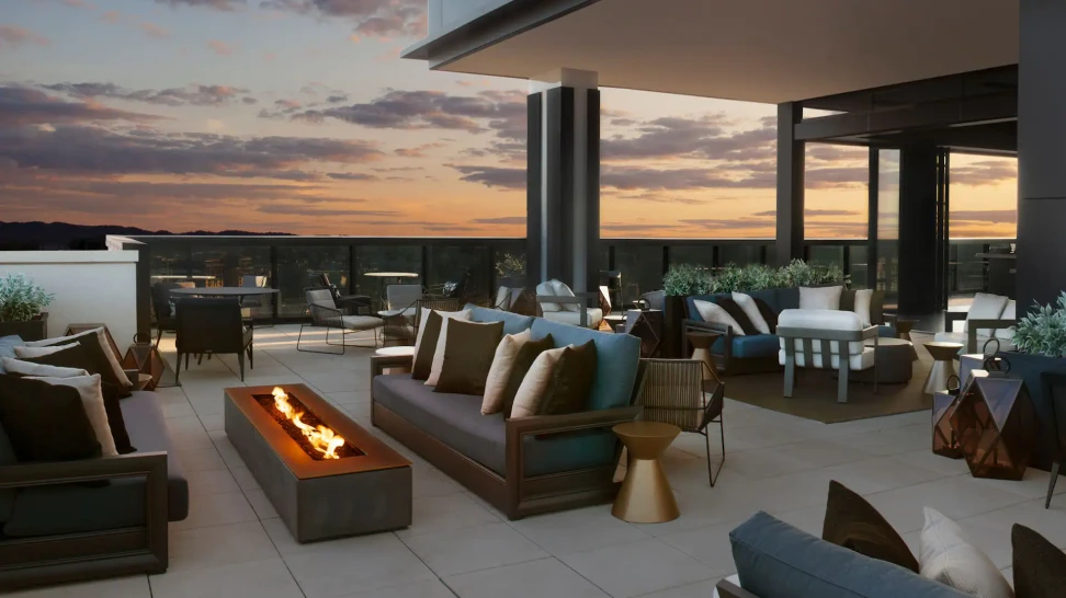 Outdoor rooftop lounge with modern seating, a fire pit, and sunset views in the background.