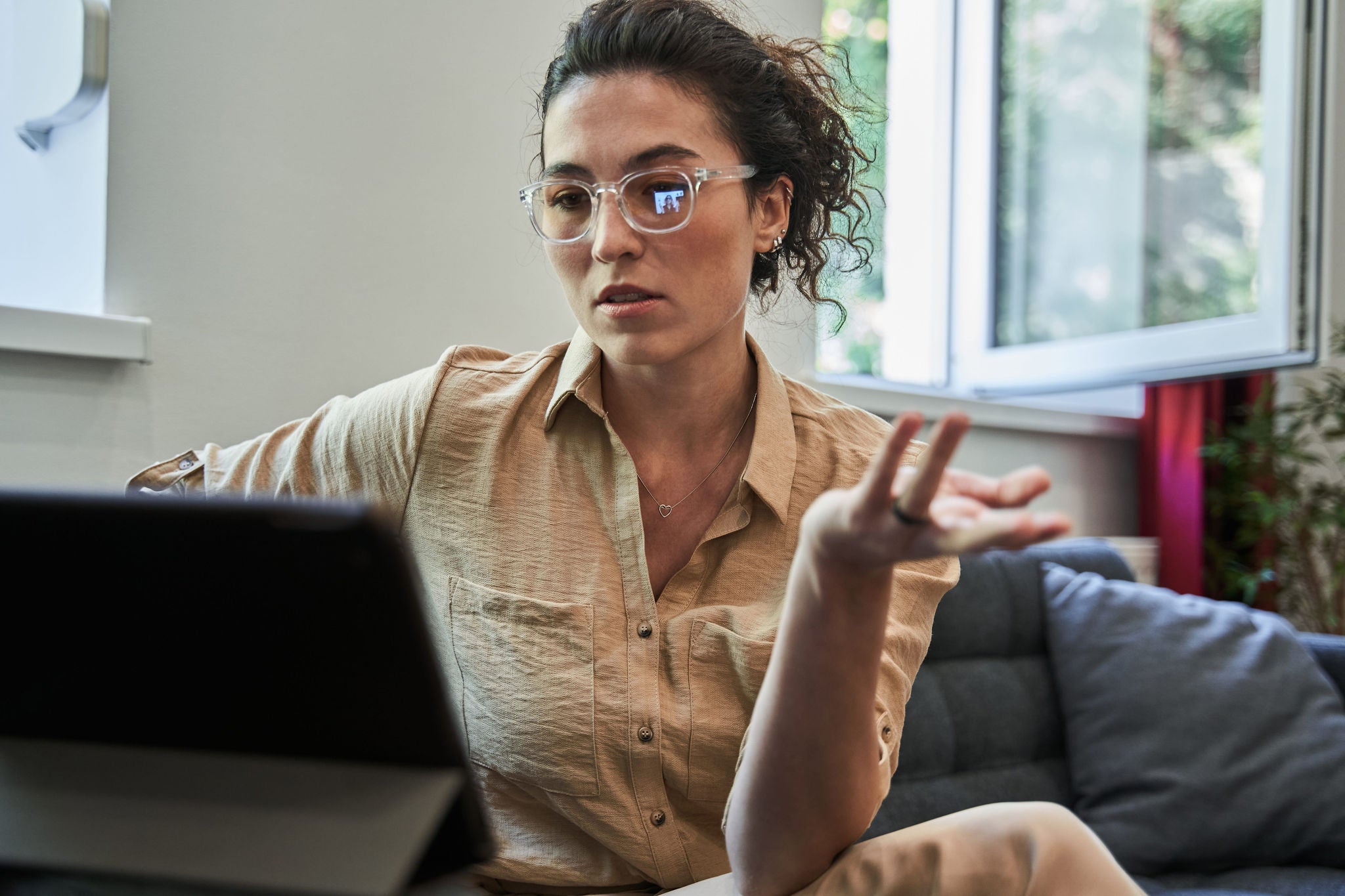 A woman with glasses sitting on a couch, gesturing while having a video therapy session on a tablet