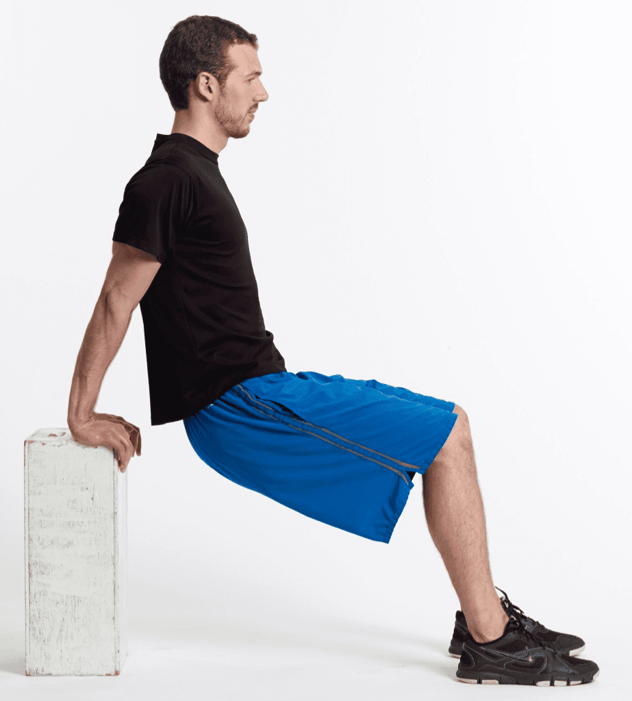 Hombre haciendo un ejercicio de tríceps utilizando un banco, con los brazos estirados y el cuerpo elevado frente al banco, vistiendo camiseta negra y pantalones cortos azules.