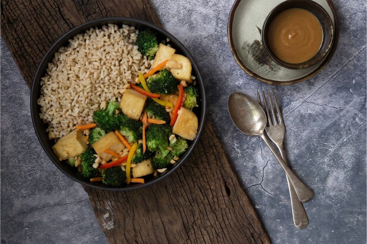 Bowl with brown rice, vegetable mixture, tofu cubes and chopped peanuts next to a small bowl of brown dressing.