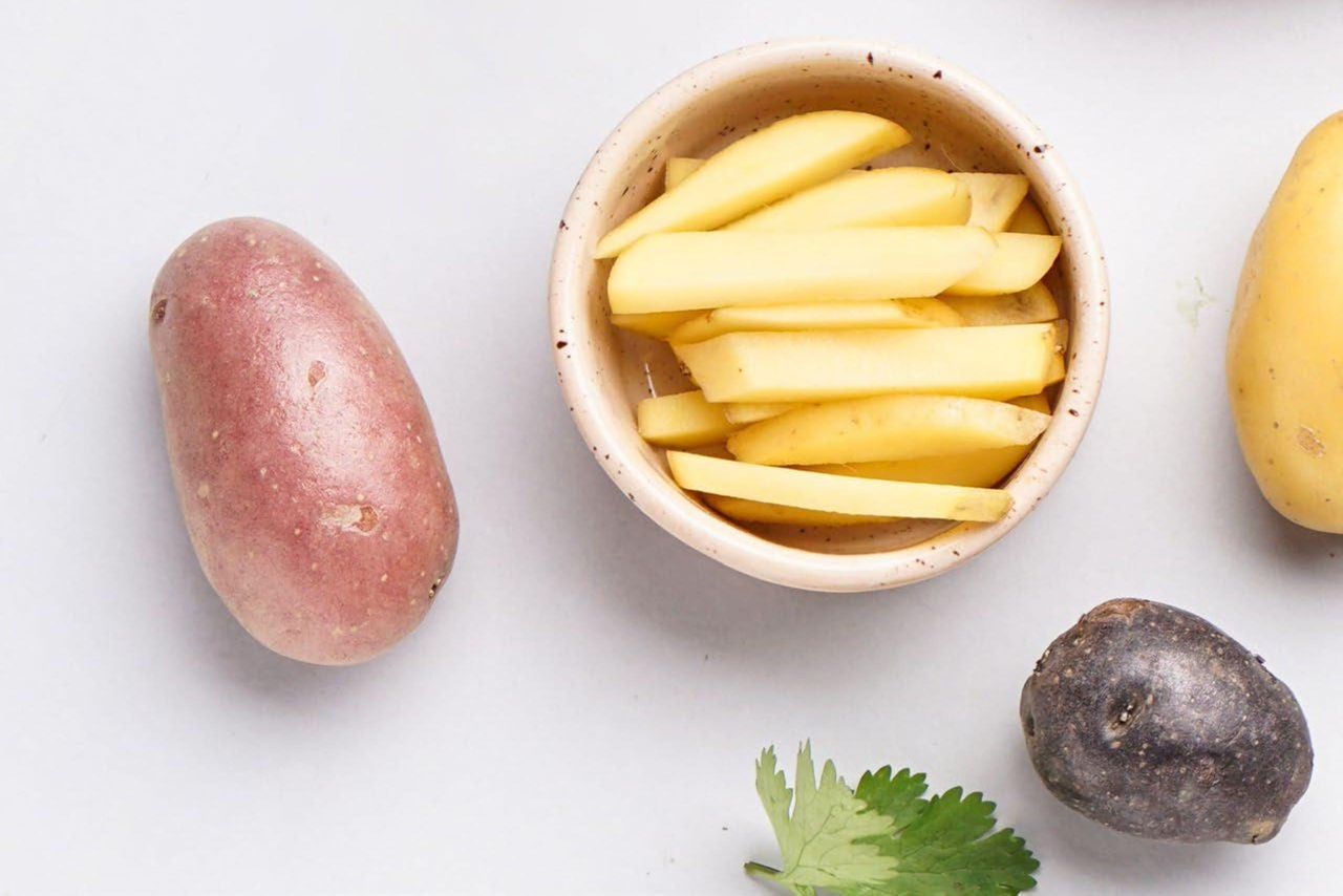 A collection of potatoes in different colors, shapes and sizes, including a potato cut into french fry shape in a bowl on a white counter.