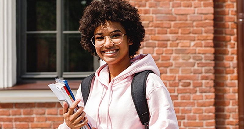 A young woman with curly hair and glasses smiling while holding books and a notebook.