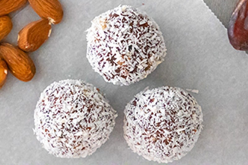 Three coconut-covered brown dessert balls on a white surface, with whole almonds nearby.