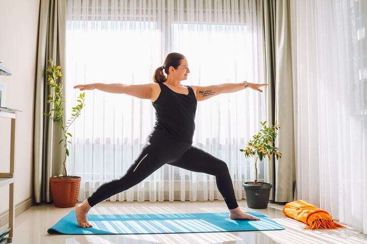 Woman smiling while doing a warrior pose in a brightly lit living room.