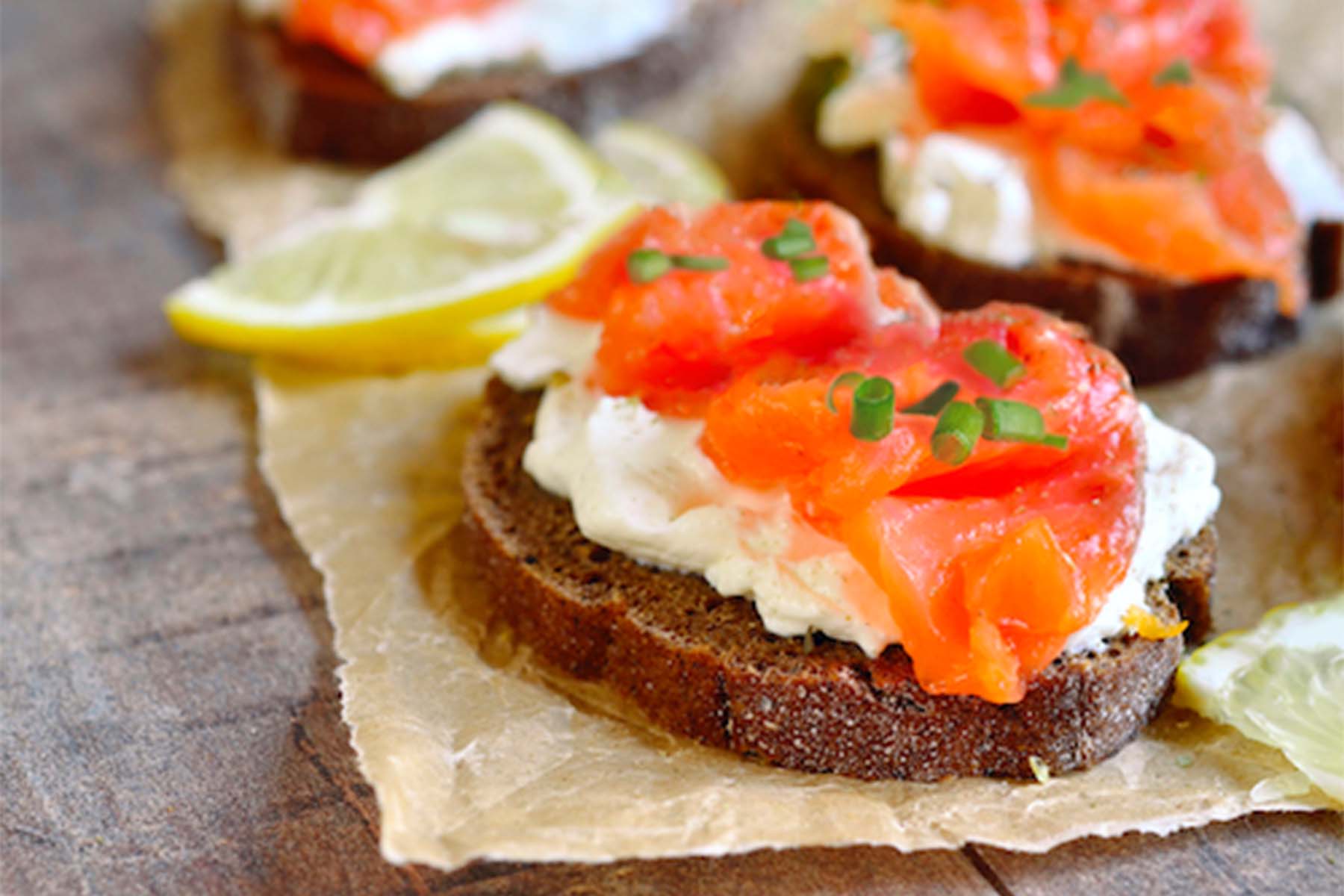 Close-up of slices of dark bread topped with cream cheese and smoked salmon, garnished with fresh herbs and lemon wedges, arranged on a rustic surface.