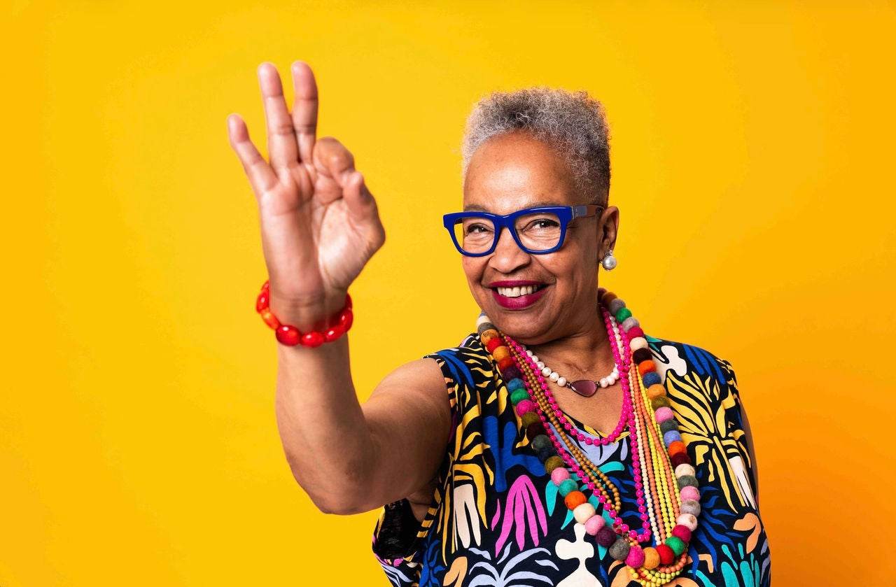 Older woman smiling while making an "okay" hand sign in front of a vibrant yellow background.