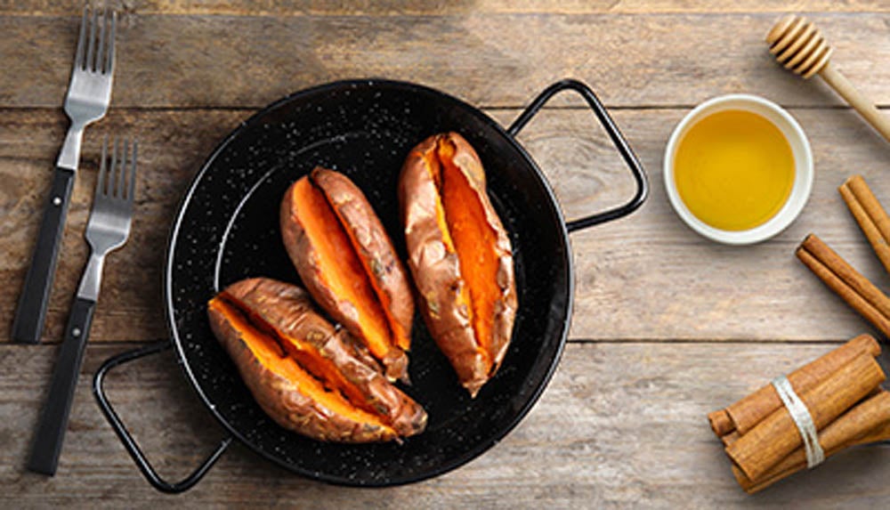 Sartén con batatas asadas acompañada de miel y ramas de canela sobre una mesa de madera.