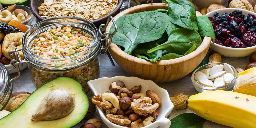 A variety of healthy foods displayed on a table, including nuts, fruits, spinach, and grains, arranged in bowls and containers.
