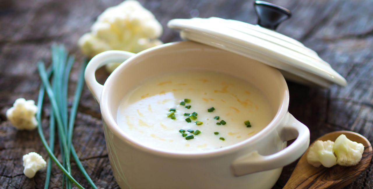 Creamy cauliflower soup garnished with chopped chives in a white ceramic bowl with a lid, set on a rustic wooden surface. Chives and cauliflower pieces are beside it.