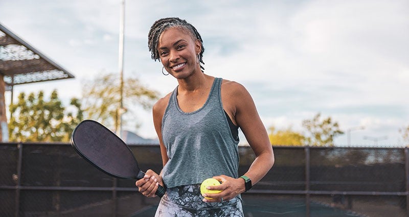 A smiling woman holding a tennis racket and a tennis ball, standing on a tennis court. She is wearing a sleeveless top and has her hair styled in a braid, with a cloudy sky in the background.
