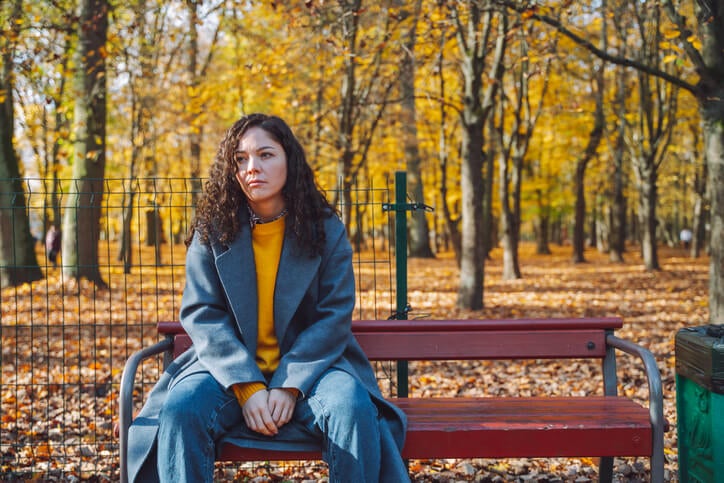 Woman with a contemplative expression sitting on a public park bench with fall foliage behind her.