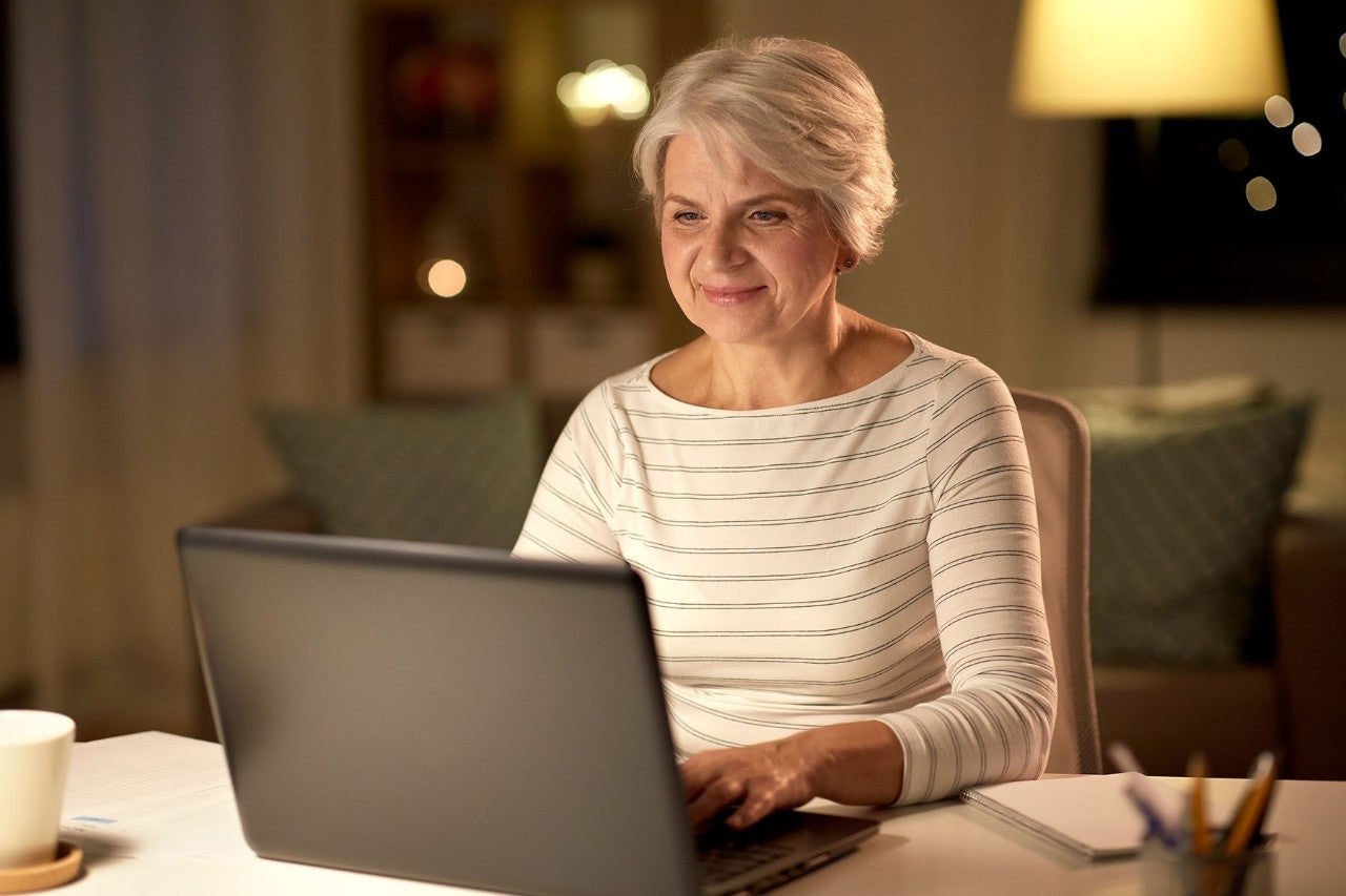 Smiling older woman using a laptop at night, with a mug and notebook nearby.