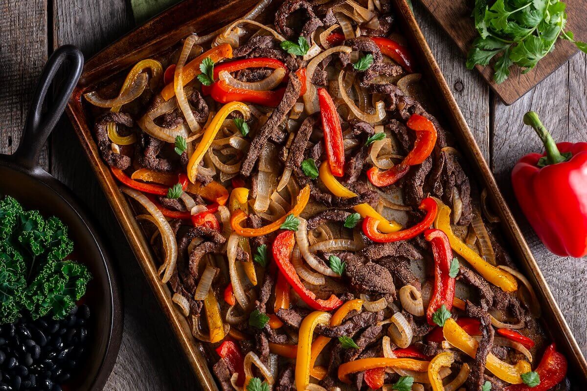 Steak and fajita vegetables on a sheet pan, next to herbs, beans and a bell pepper.