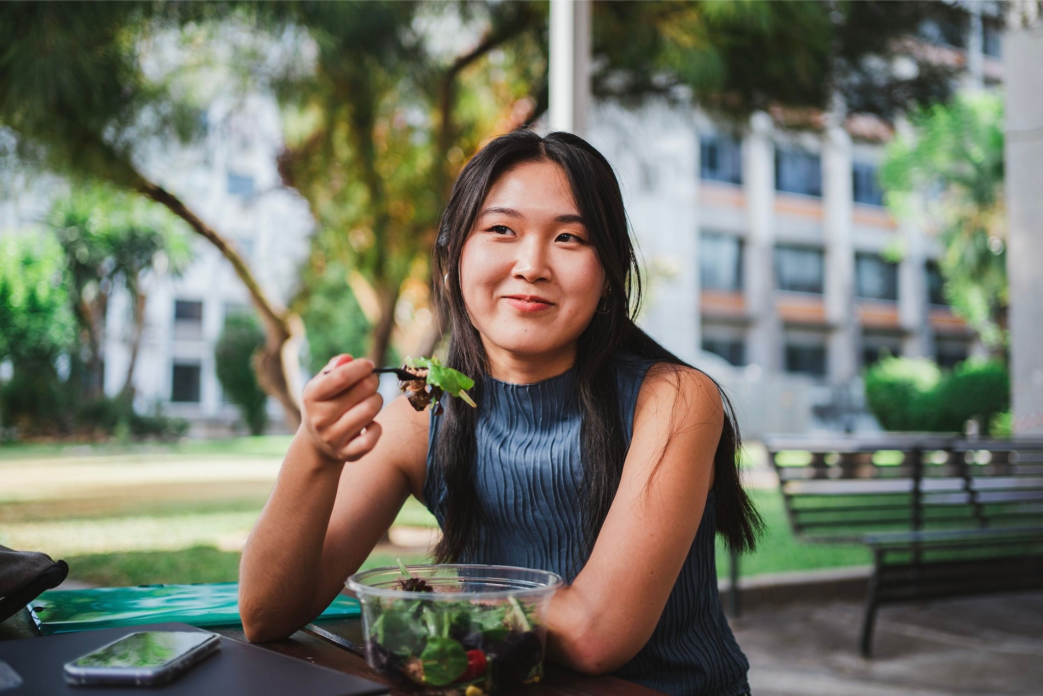 Woman sitting at a table in a park eating salad while smiling.