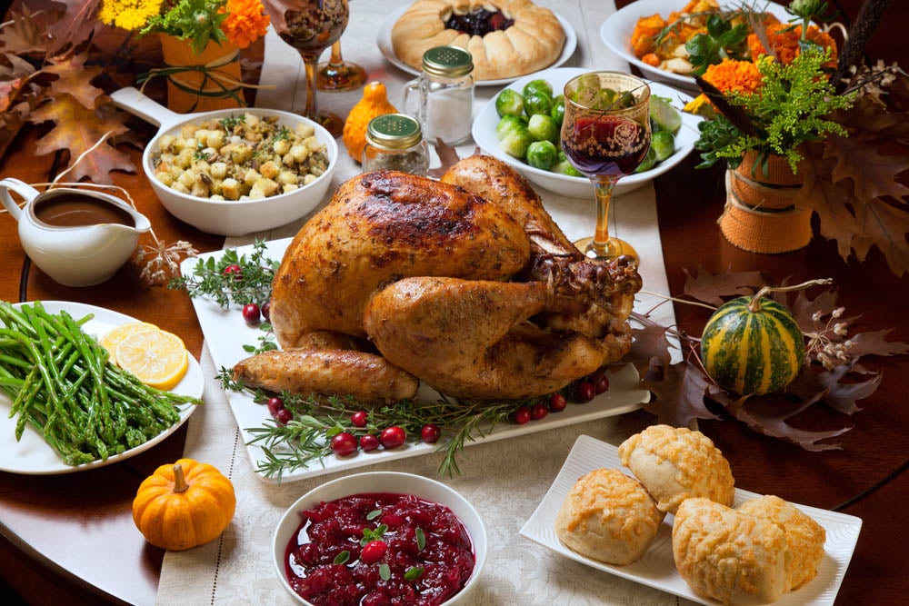 A Thanksgiving table with a roasted turkey, cranberries, stuffing, vegetables, rolls, pie, and decorative gourds, surrounded by autumn leaves.