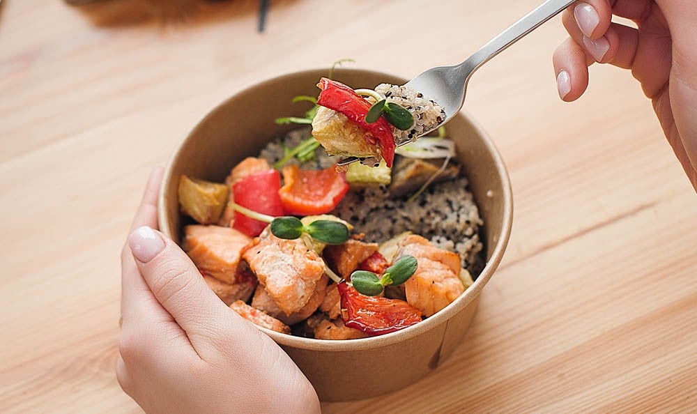 A person's hand holding a fork above a bowl filled with salmon, vegetables, and rice, placed on a wooden table.