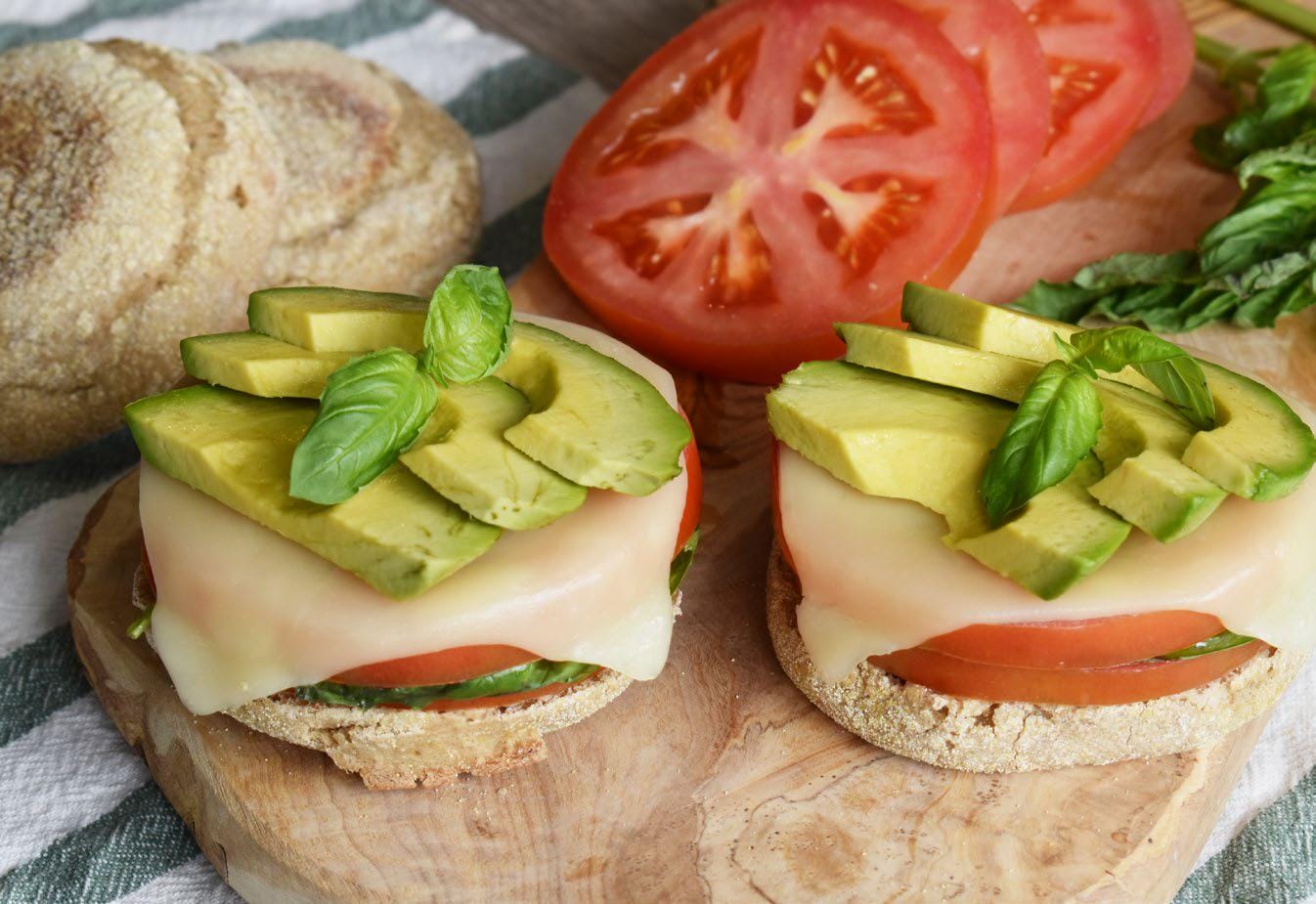 Open-faced sandwich with melted cheese, sliced avocado, tomato, and fresh basil on an English muffin. Sliced tomatoes and basil leaves are in the background.