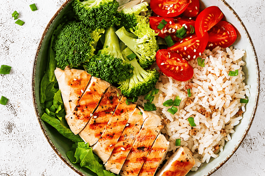 White bowl with sliced grilled chicken, broccoli, and tomatoes on rice sitting on a white counter.