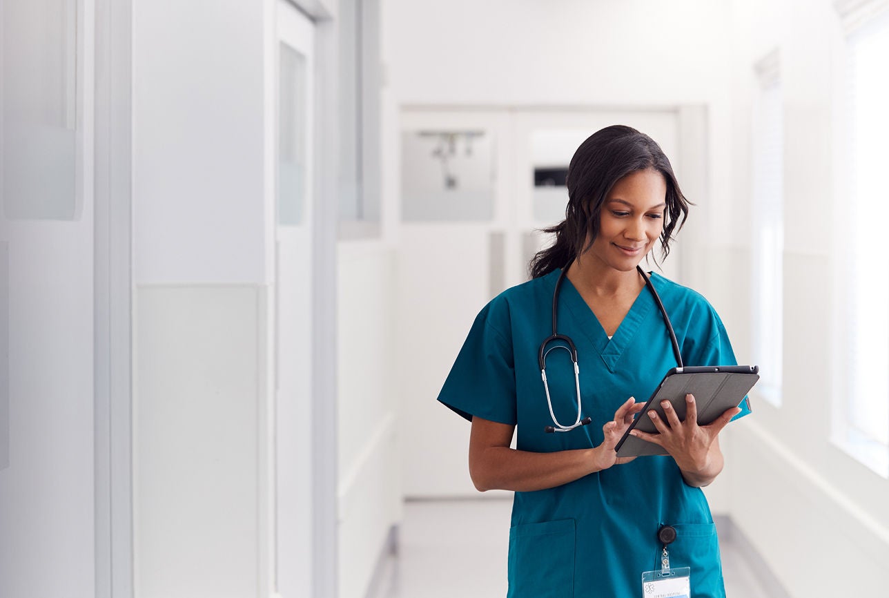 Nurse in teal scrubs using a tablet while walking through a bright hospital corridor