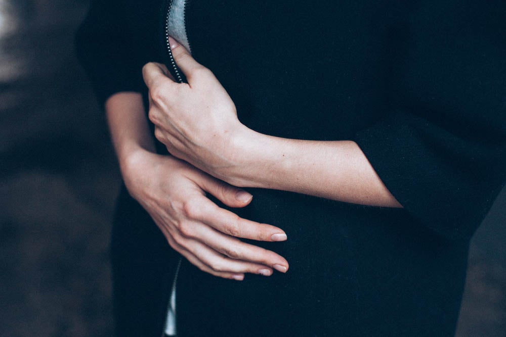 Person in a dark sweatshirt with hands held over their stomach, standing against a blurred background.
