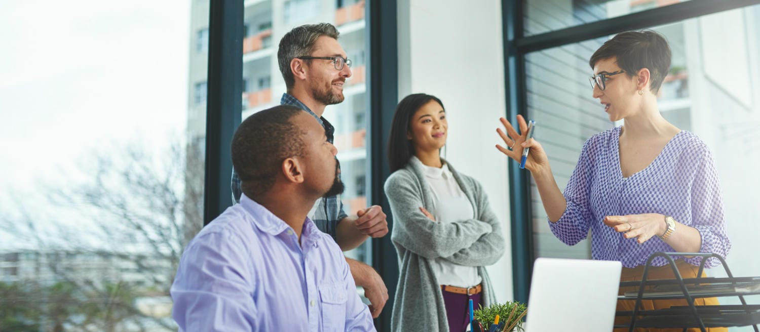 A group of four people in a modern office, with one woman speaking while others listen attentively