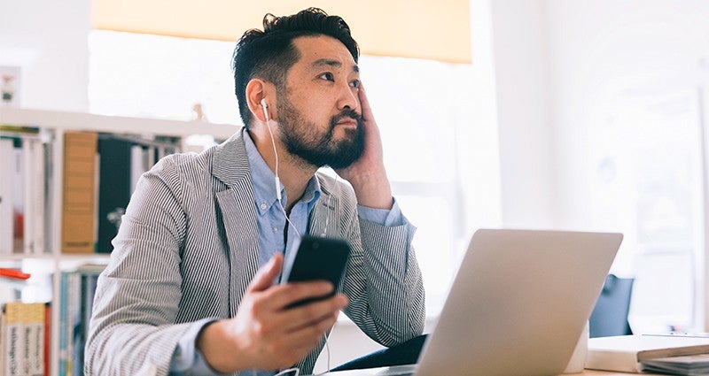 Man in a striped blazer sitting at a desk, holding a smartphone and listening with earphones while working on a laptop