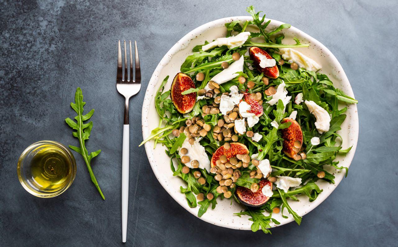 A fresh salad with arugula, figs, cheese, and lentils is served on a white plate. A fork and a small glass of olive oil are placed beside it on a gray surface.