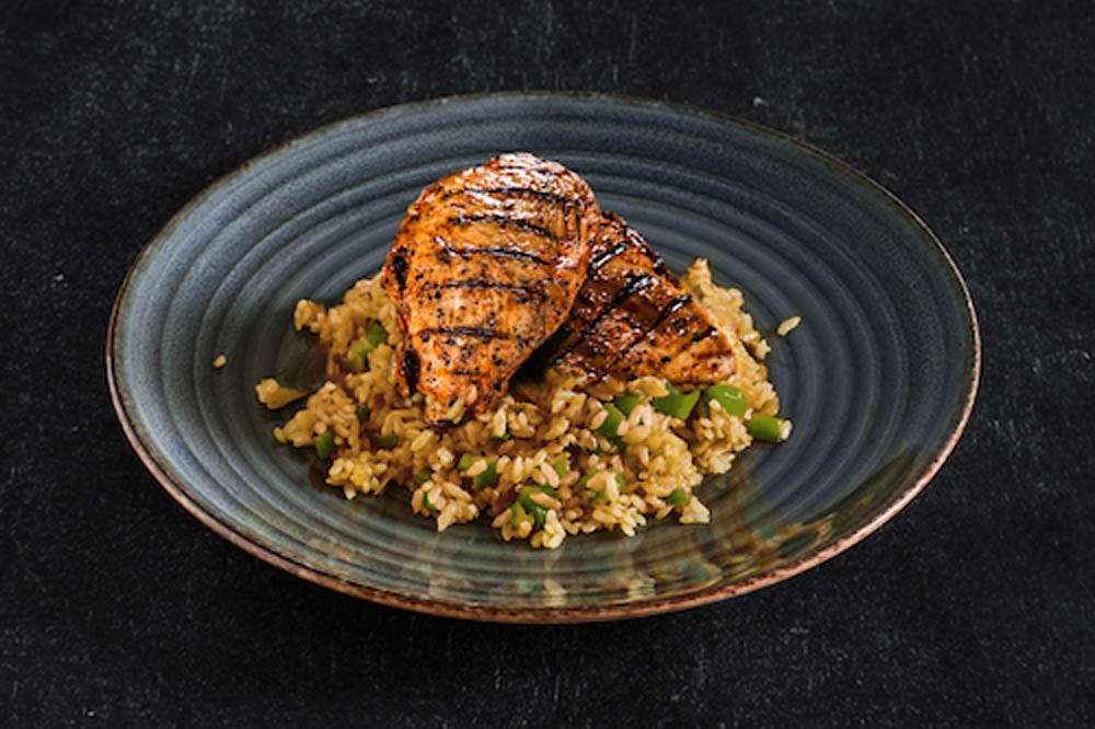 Pechuga de pollo a la parrilla servida sobre una cama de arroz frito con verduras en un plato de cerámica oscuro