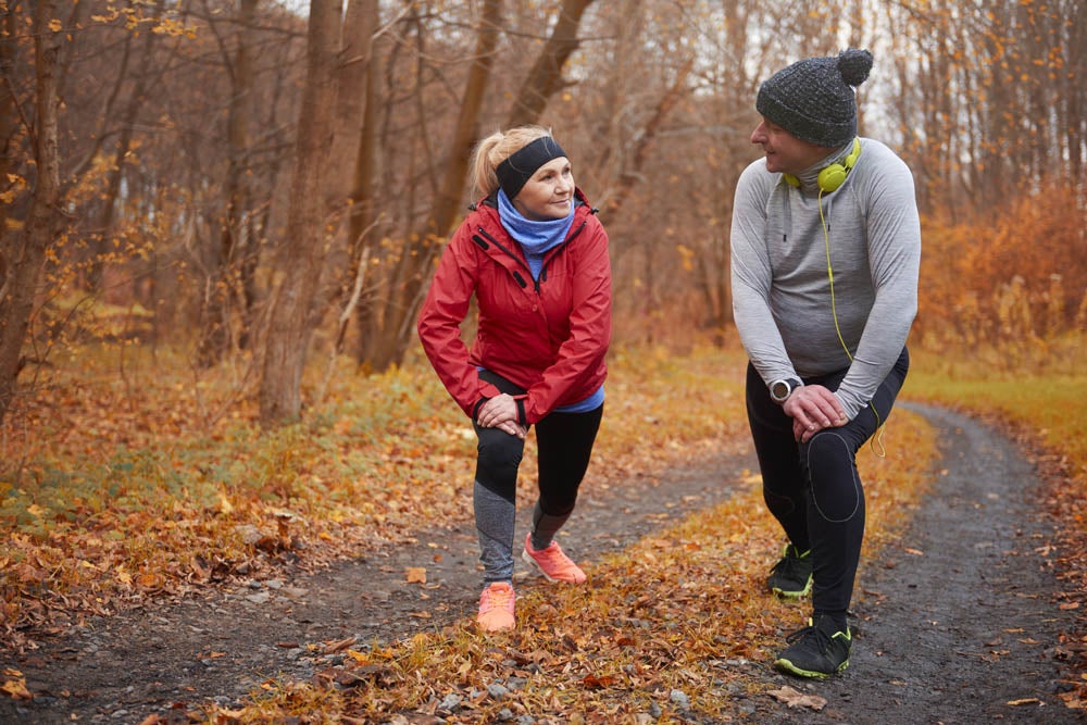 Two people stretching and lunging on a leaf-covered path in a forest during autumn. Both are wearing athletic gear and are engaged in conversation.