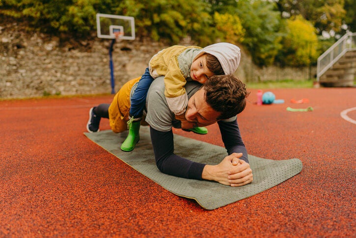 Man performing a plank exercise on a yoga mat in a park with a small child on his back. 