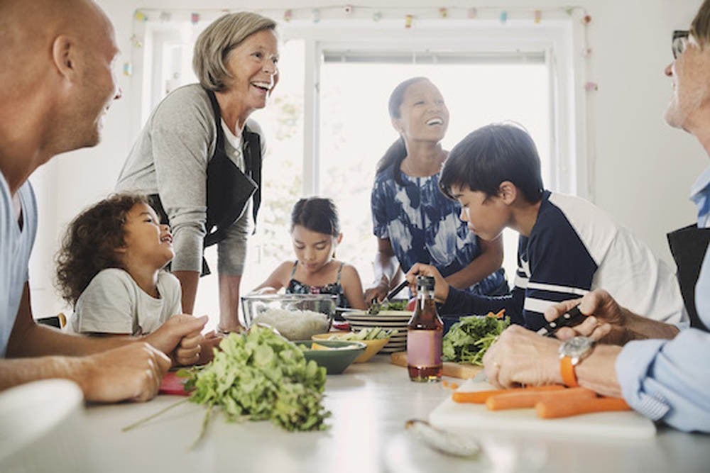 A diverse group of people, including adults and children, gather around a table with fresh vegetables, smiling and preparing food together.