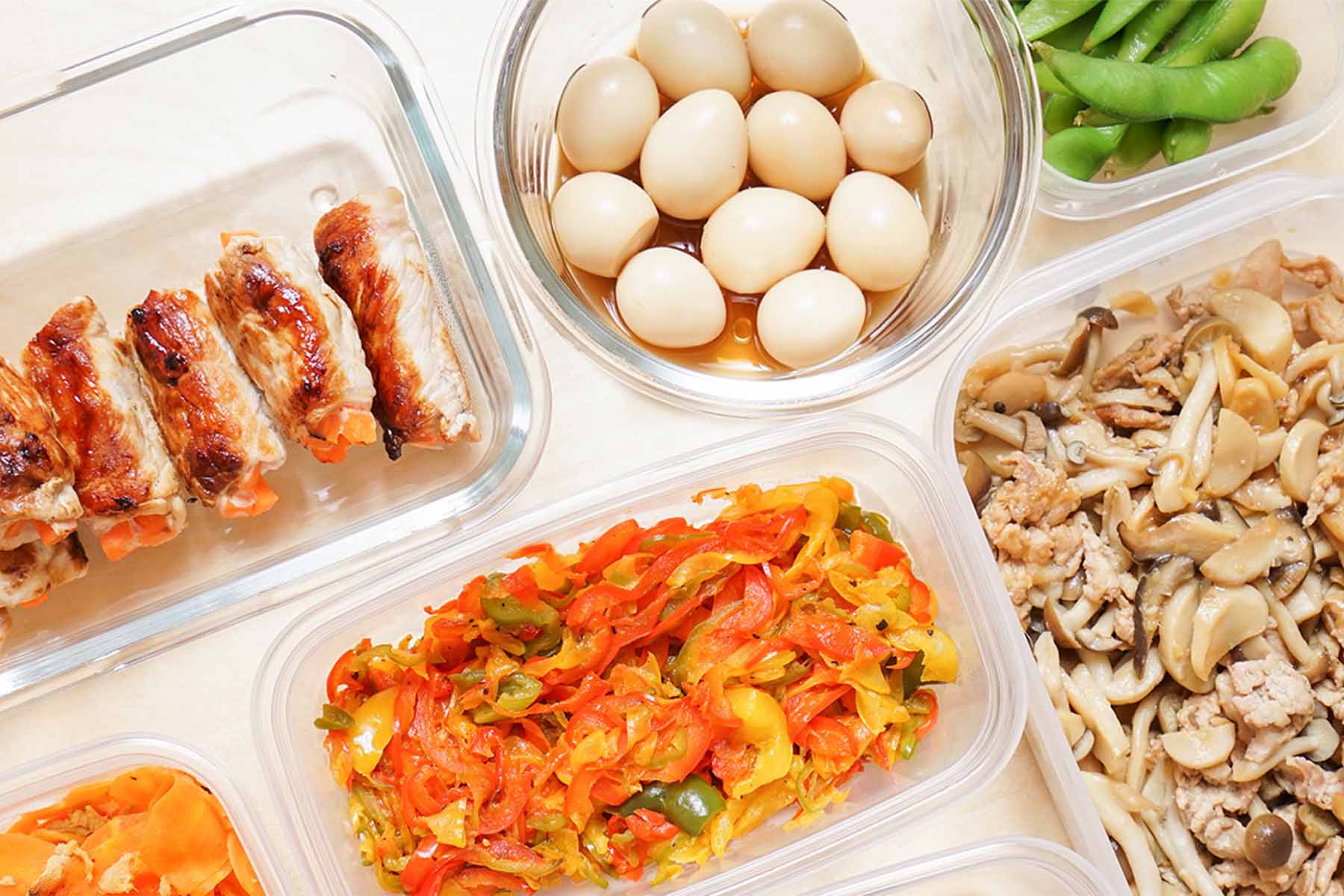 Variety of meal prep containers filled with grilled chicken rolls, boiled eggs, green edamame, sautéed vegetables, and mushrooms with ground meat, arranged on a white surface.