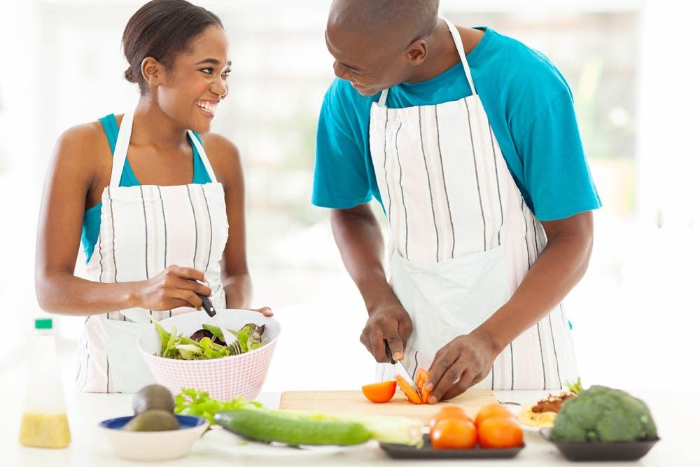 Pareja sonriendo mientras prepara una ensalada juntos en la cocina, usando delantales rayados