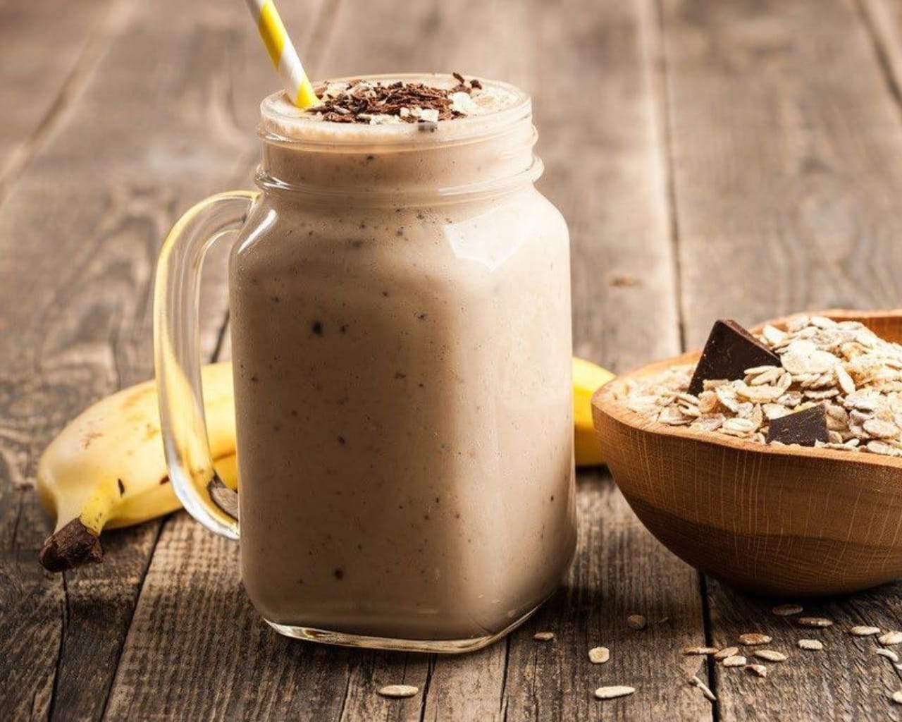 A creamy chocolate banana protein smoothie in a large glass mug, garnished with chocolate shavings on top with a banana behind and next to a bowl with ingredients in it on a wood table.