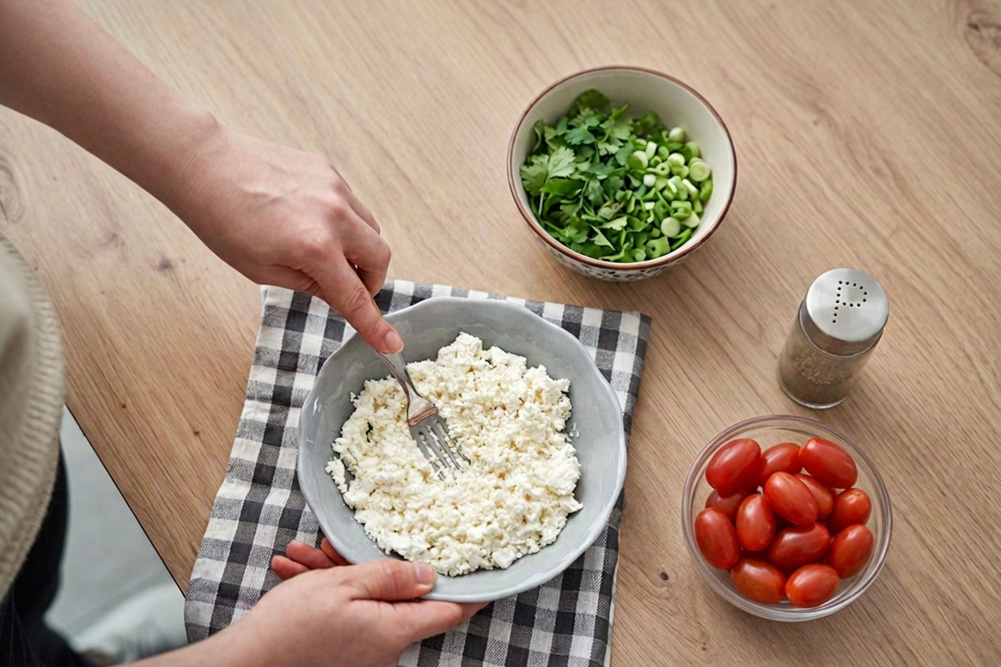 Overhead view of a person scooping cottage cheese into a bowl next to bowls of grape tomatoes and chopped herbs.