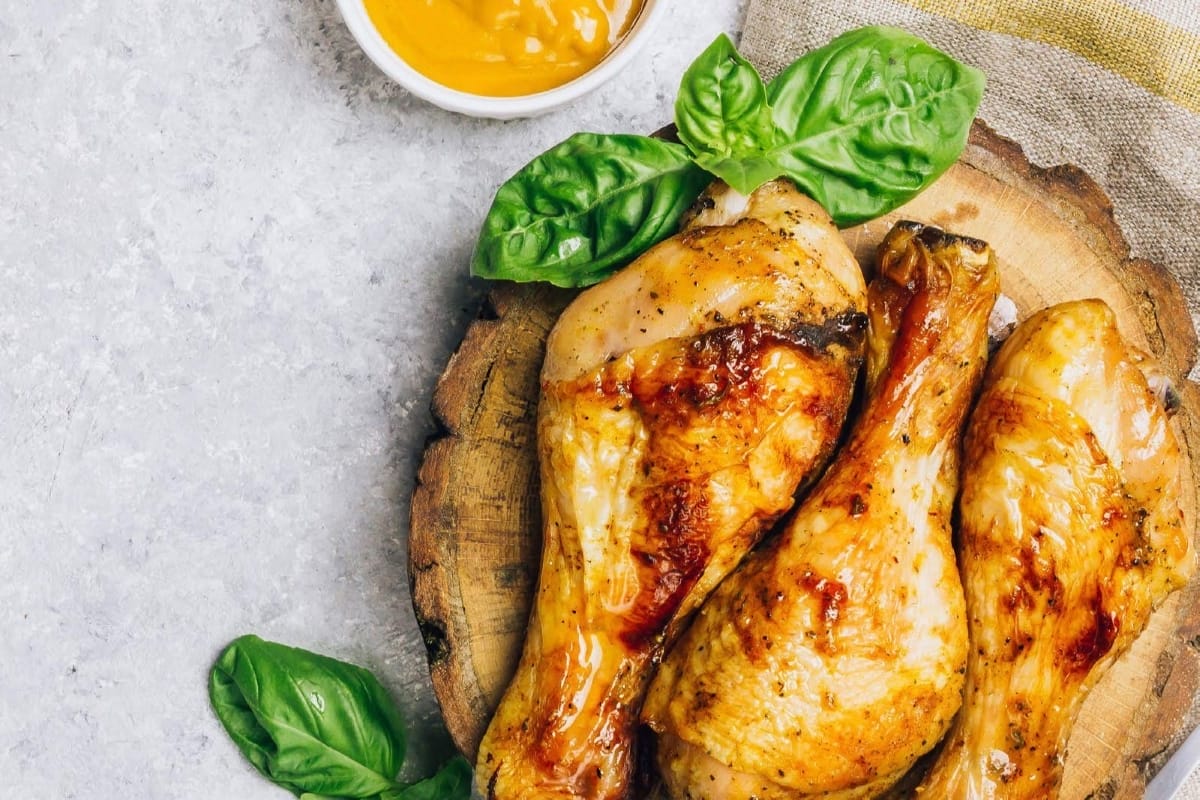 Chicken legs on a round wood block with green leaves as garnish and sauce in the background on a light grey counter.